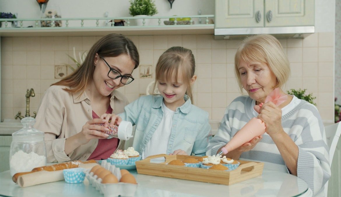 Child, mother and grandmother are decorating cupcakes and talking in kitchen enjoying leisure time together. People, children and culinary concept.